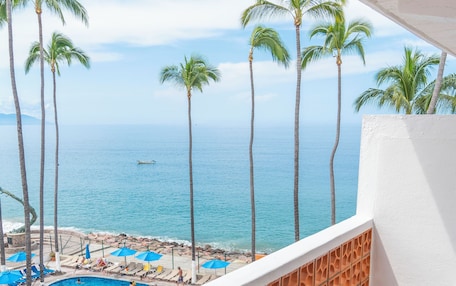 A balcony with wicker chairs, a pool, and a view of the ocean and palm trees.