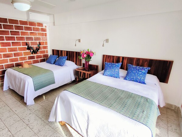 Habitación de hotel con dos camas, cada una con cabecera de madera, ropa de cama blanca y almohadas azules. Se aprecia una pared de ladrillo, una mesita con un florero y un ventilador de techo.