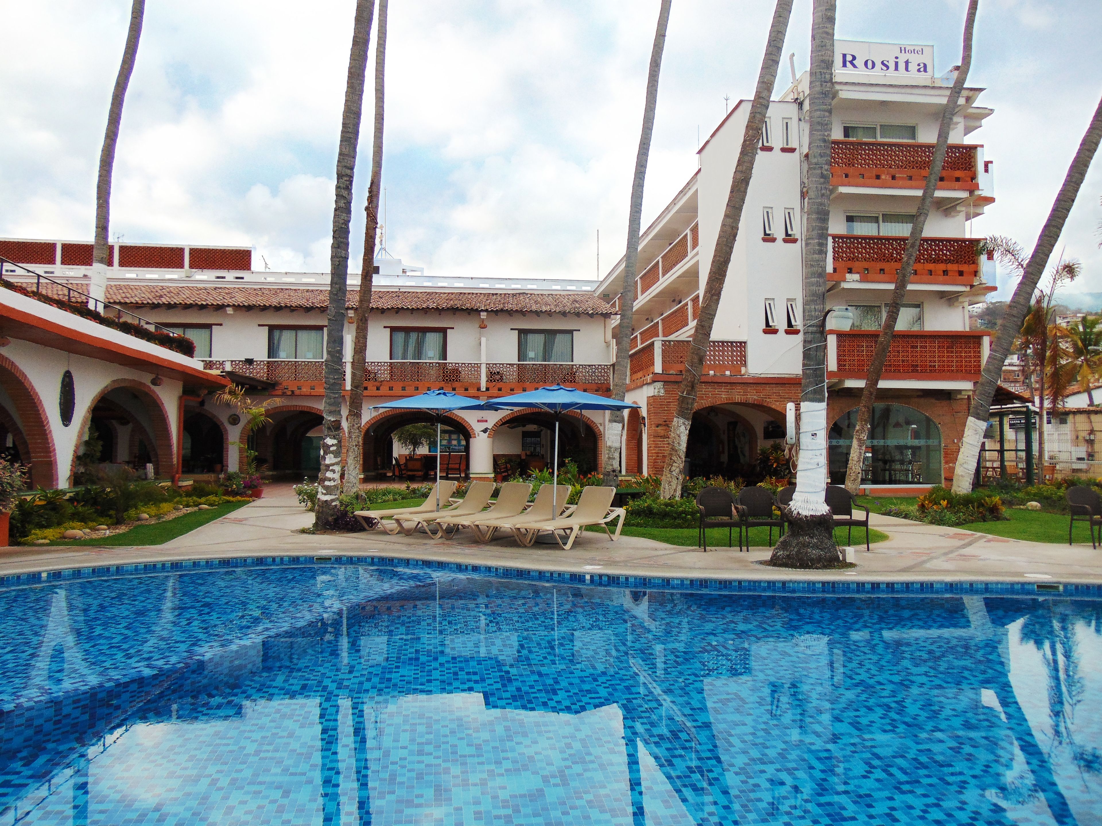 A hotel with a swimming pool, sun loungers, and palm trees.