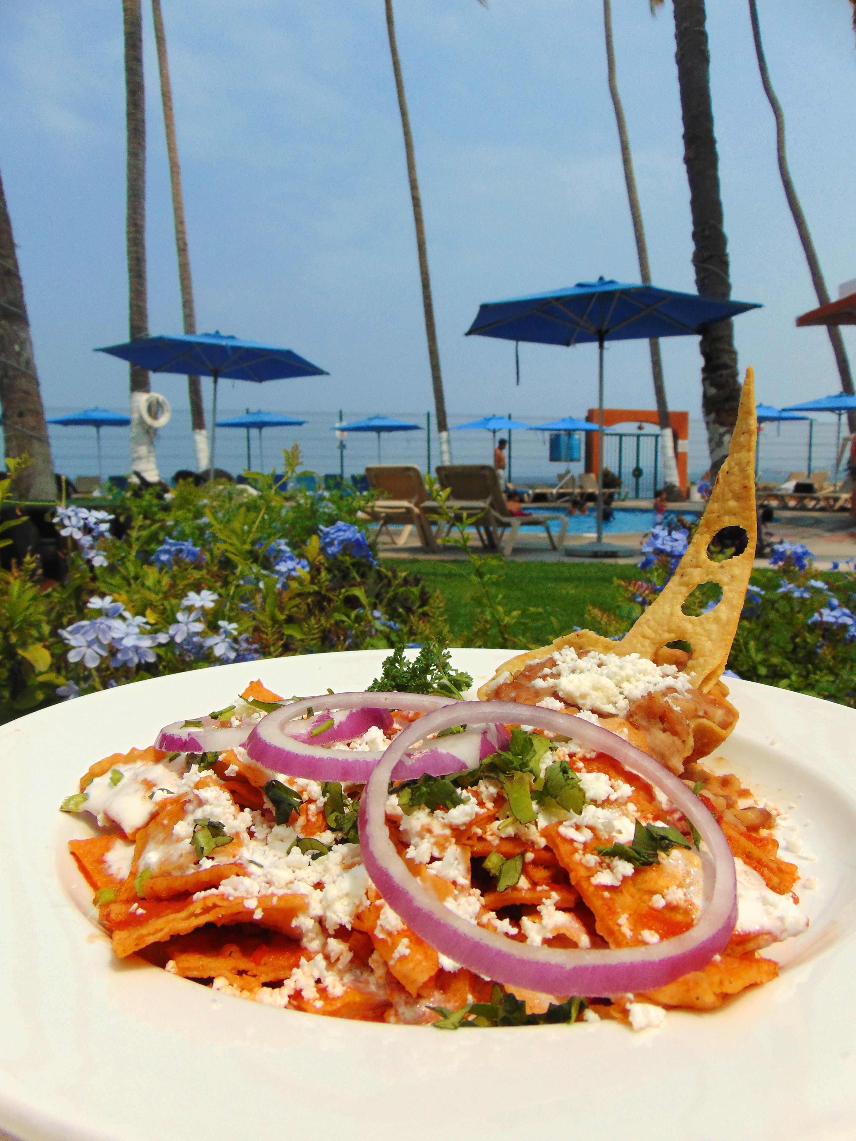 Une assiette de nachos garnis, placée sur fond de plage avec des palmiers et des parasols.