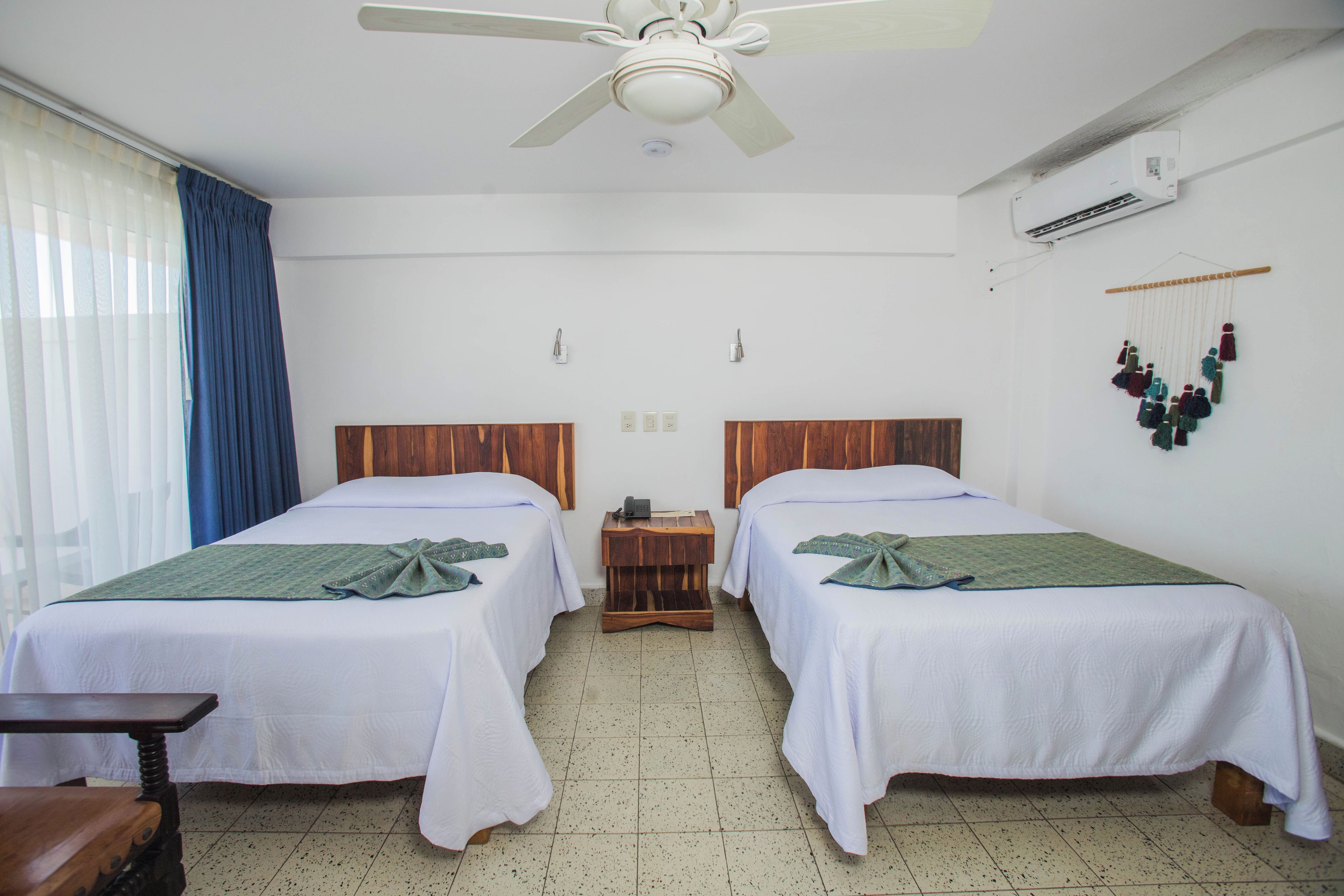 Een hotelkamer met twee bedden, een plafondventilator, een airconditioningsapparaat en een klein houten nachtkastje.