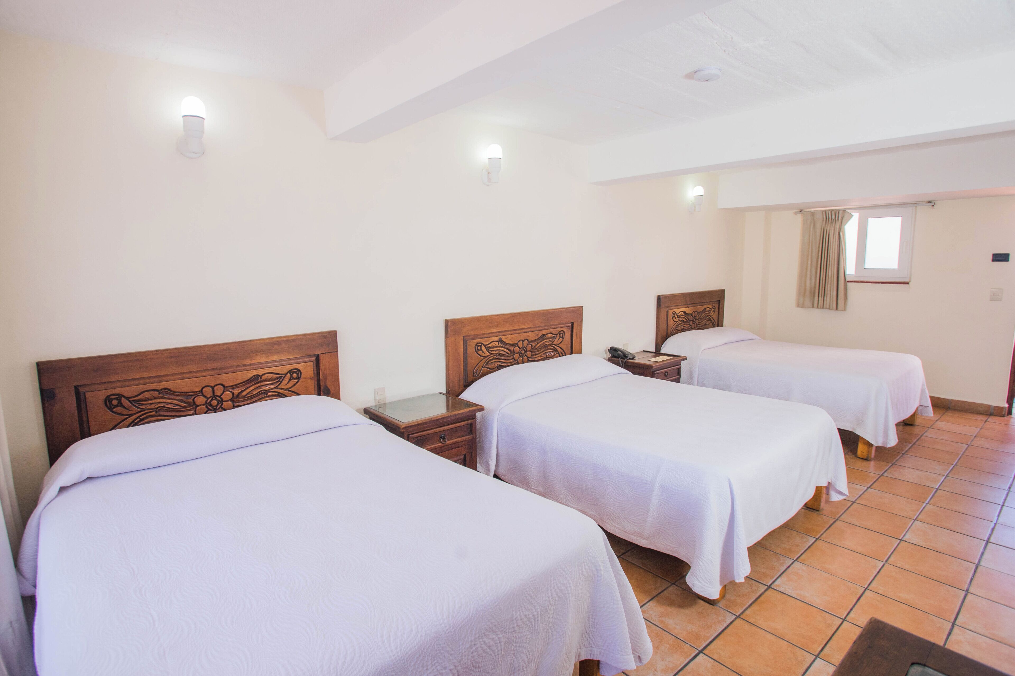Una habitación de hotel con tres camas individuales, cabeceros de madera, mesitas de noche y una ventana con cortinas.