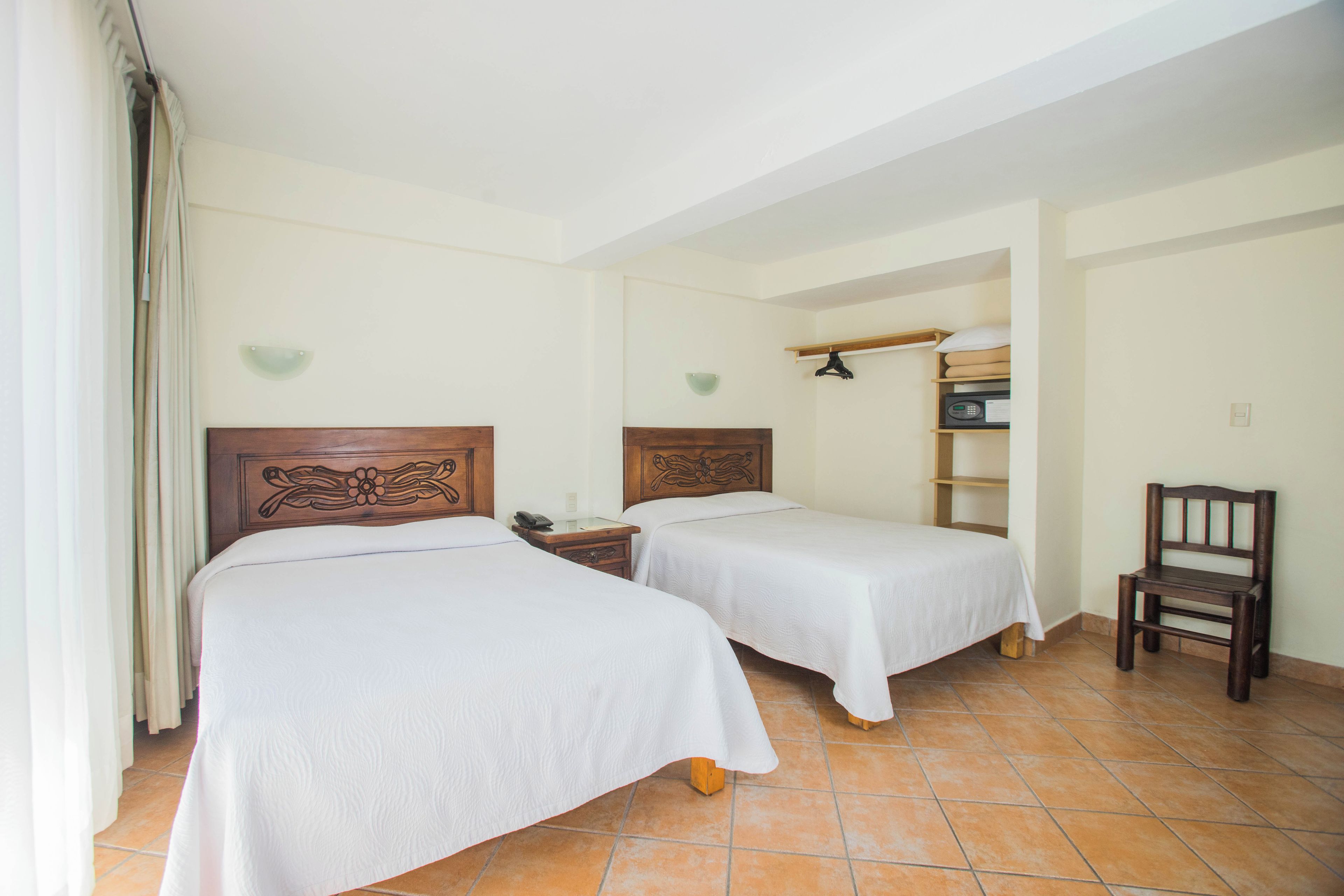 A hotel room with two single beds, a bedside table with a phone, a small shelf with a pillow, and a chair.