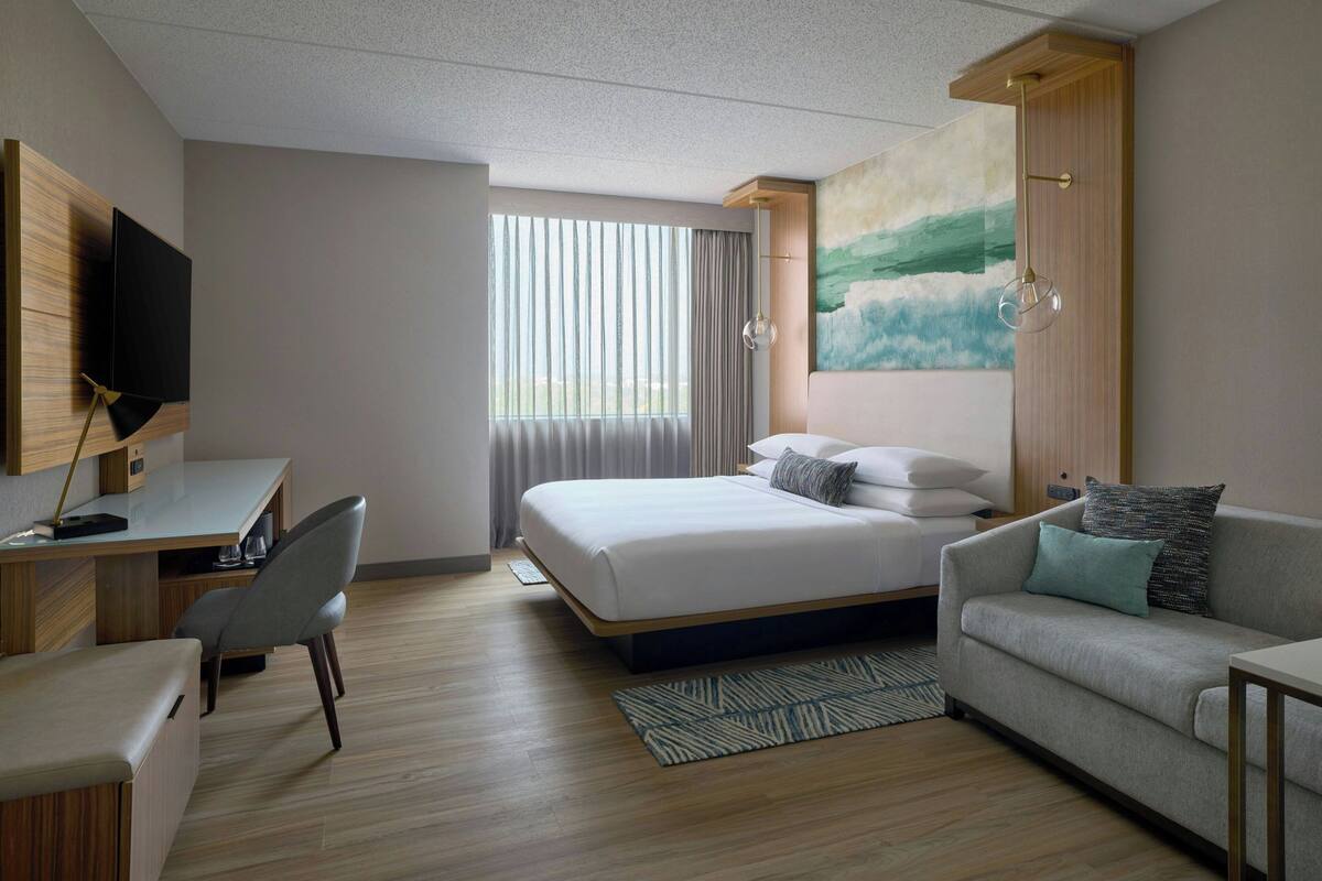 A modern hotel room with a bed, a sofa, a desk with a chair, a TV, and a painting on the wall.