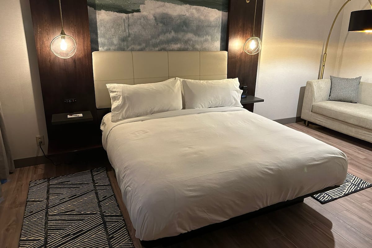 A neatly made bed with white linens, a modern abstract painting on the wall, and a side table with a lamp.