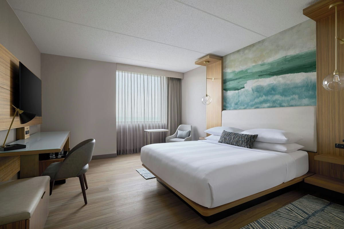 A modern hotel room with a large bed, a desk, a chair, a TV, and a painting on the wall.