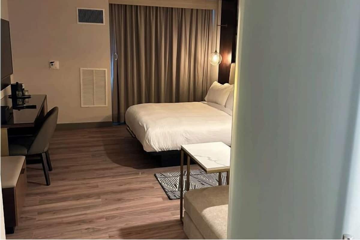 A hotel room with a bed, a desk, a chair, a bedside table, and a lamp.