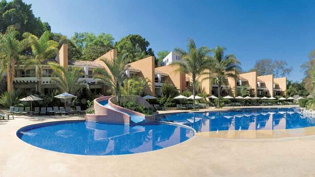 A resort with a large swimming pool, palm trees, and multiple buildings with balconies. - Hotel Rancho San Diego Grand Spa Resort (Ixtapan de la Sal)