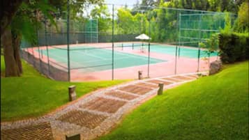 A tennis court with a surrounding fence, a pathway leading to the court, and trees in the background.
