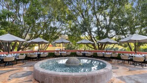An outdoor patio with a circular water feature, surrounded by tables and chairs under umbrellas, set against a backdrop of trees. - Hotel Rancho San Diego Grand Spa Resort (Ixtapan de la Sal)