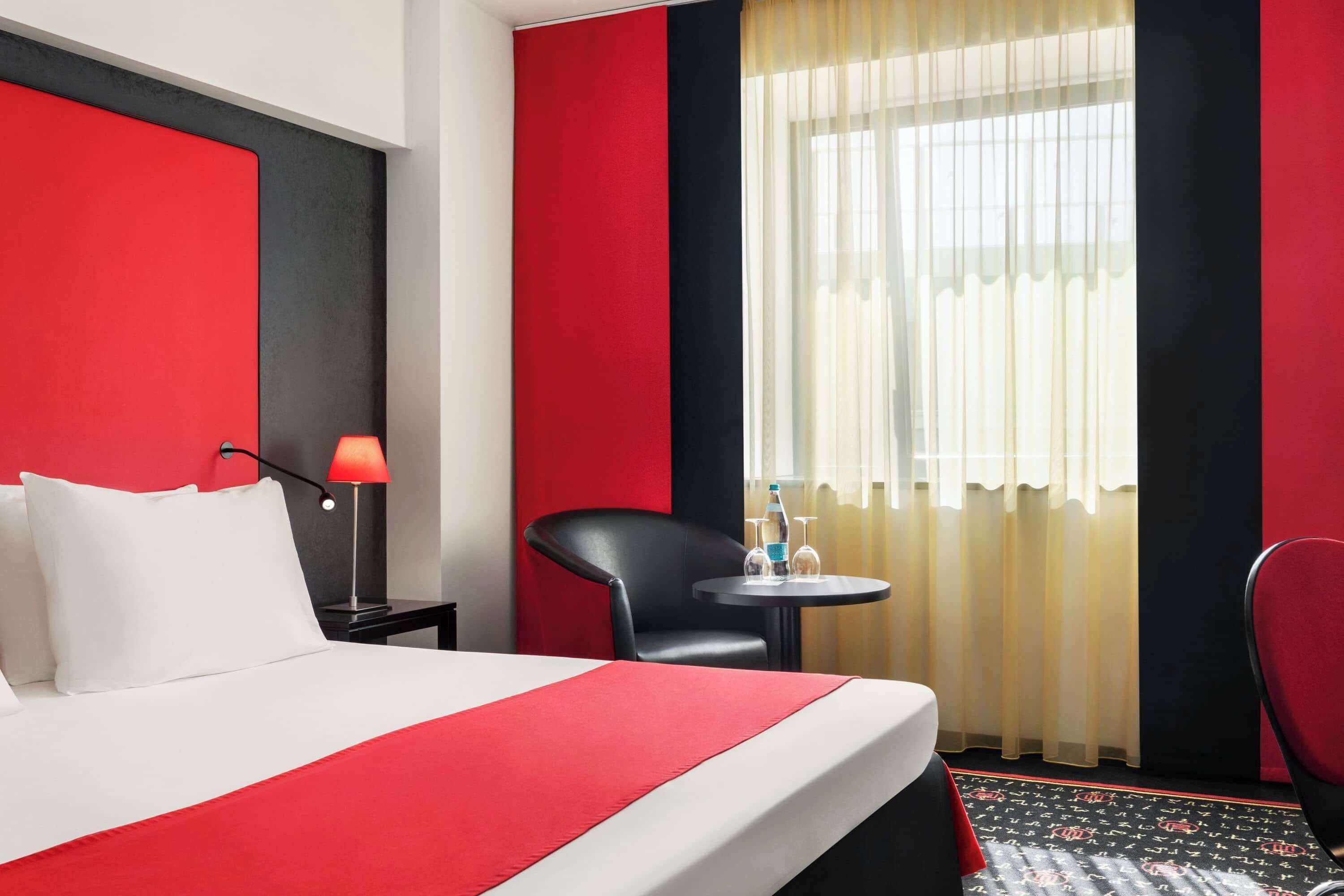 Una habitación de hotel con una cama, una mesita de noche, una lámpara, una silla, una pequeña mesa y un ventanal con cortinas.