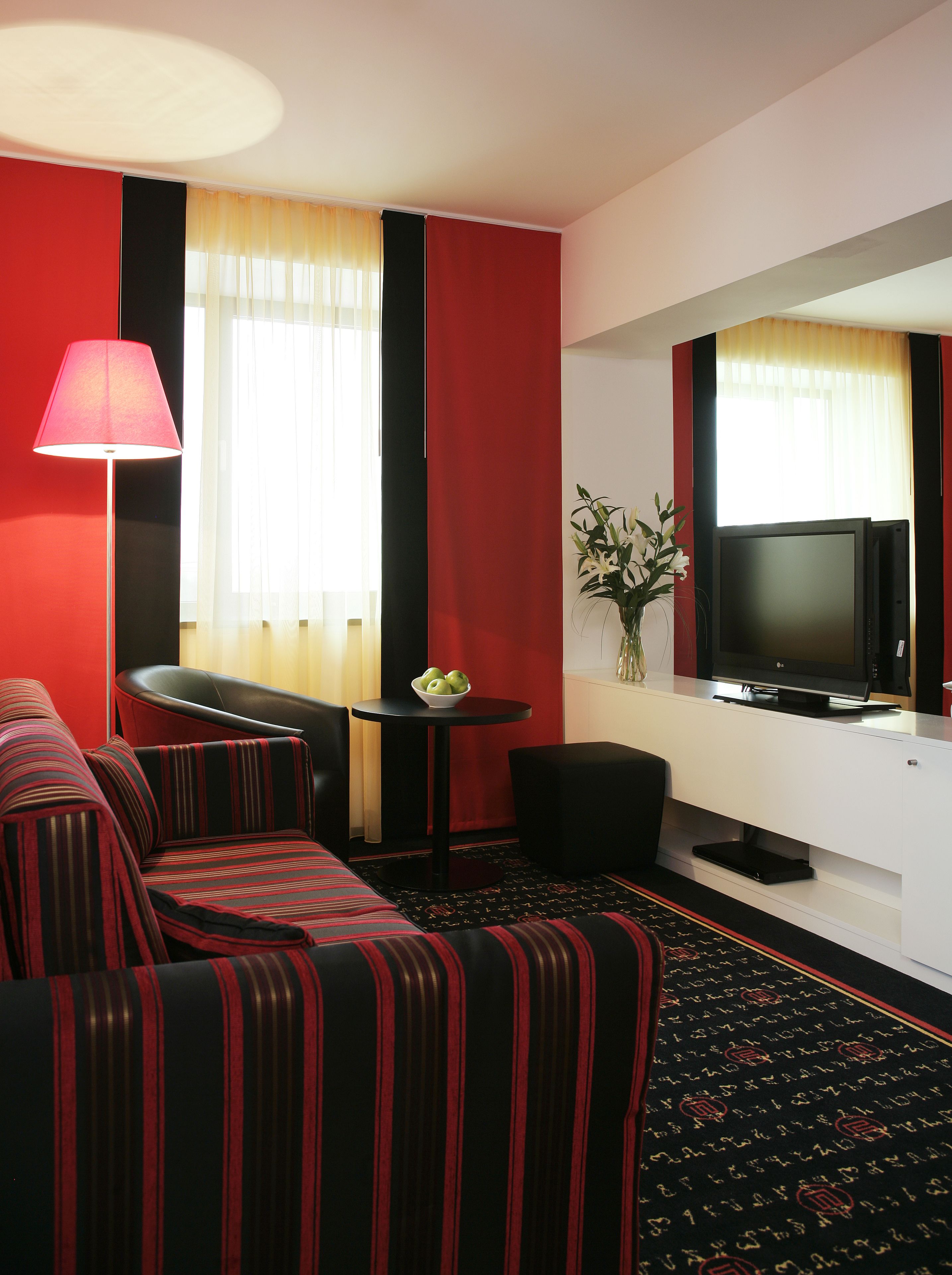 Habitación de hotel con un sofá a rayas rojo y negro, una pequeña mesa redonda con un frutero, un televisor de pantalla plana y un jarrón con flores.