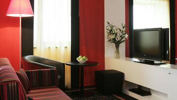 Habitación de hotel con un sofá a rayas rojo y negro, una pequeña mesa redonda con un frutero, un televisor de pantalla plana y un jarrón con flores.