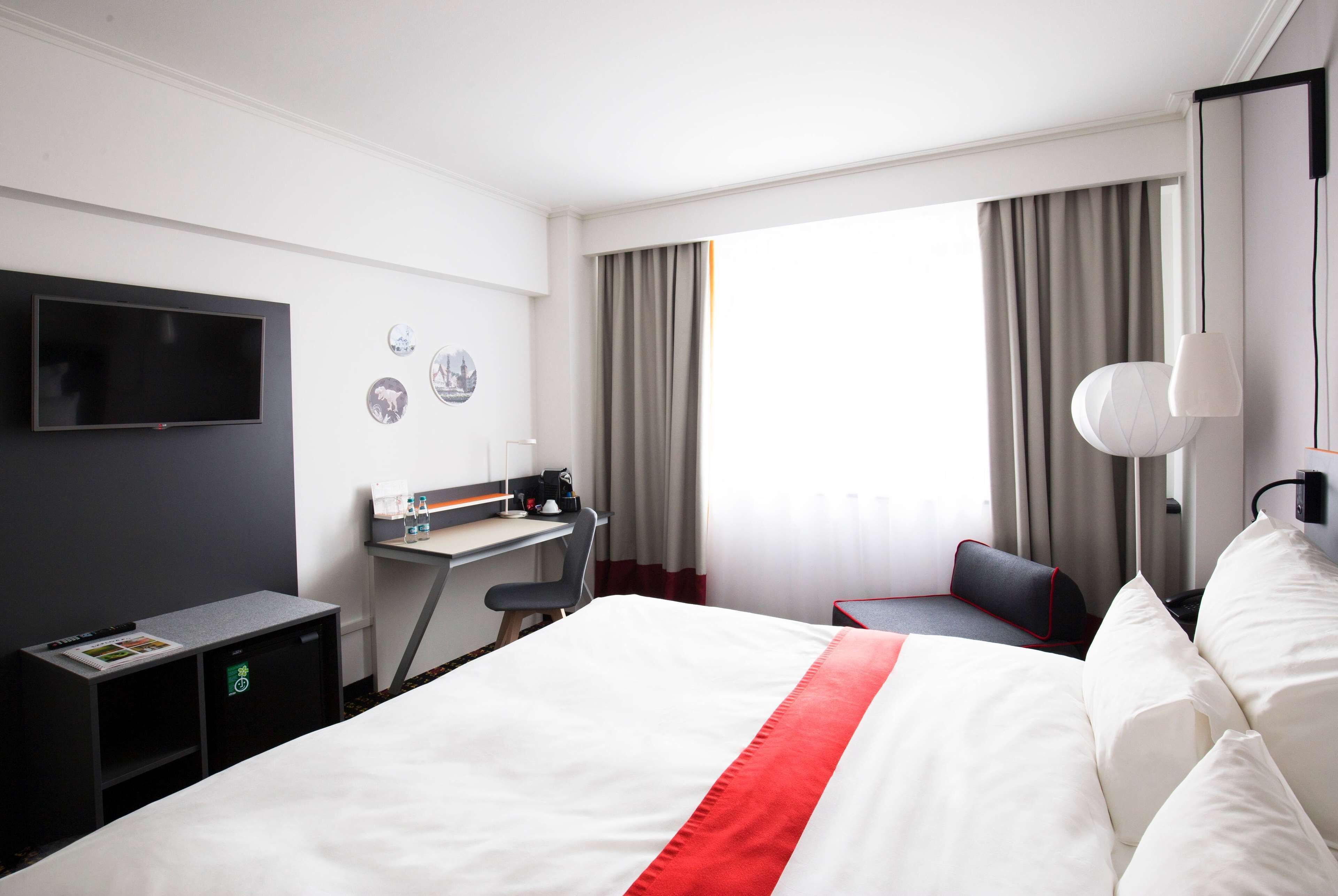 A hotel room with a large bed, a desk with a chair, a flat-screen TV mounted on the wall, a small table with a lamp, and a window with curtains.