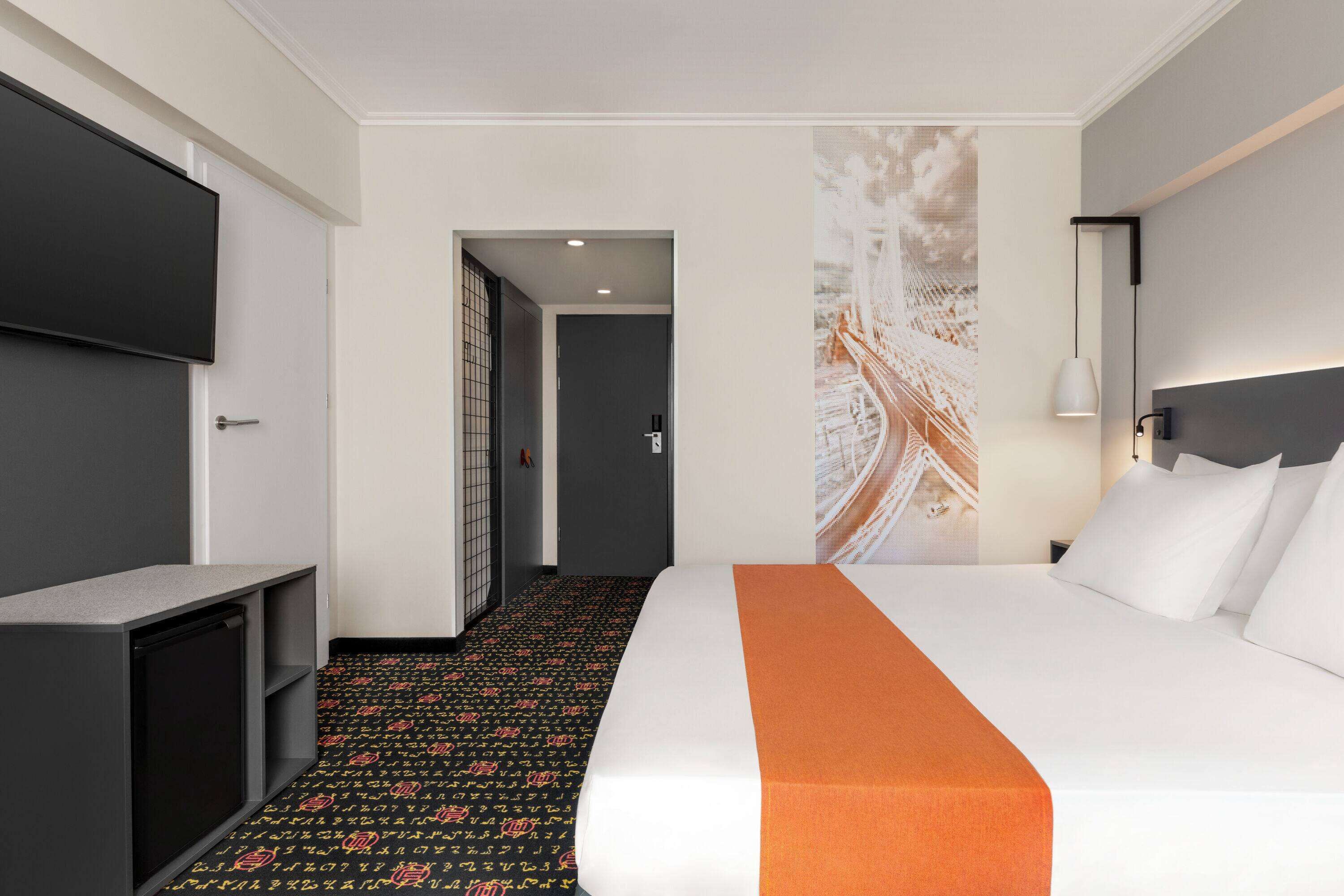 Una habitación de hotel con cama, escritorio, televisor y puerta que da a un pasillo.