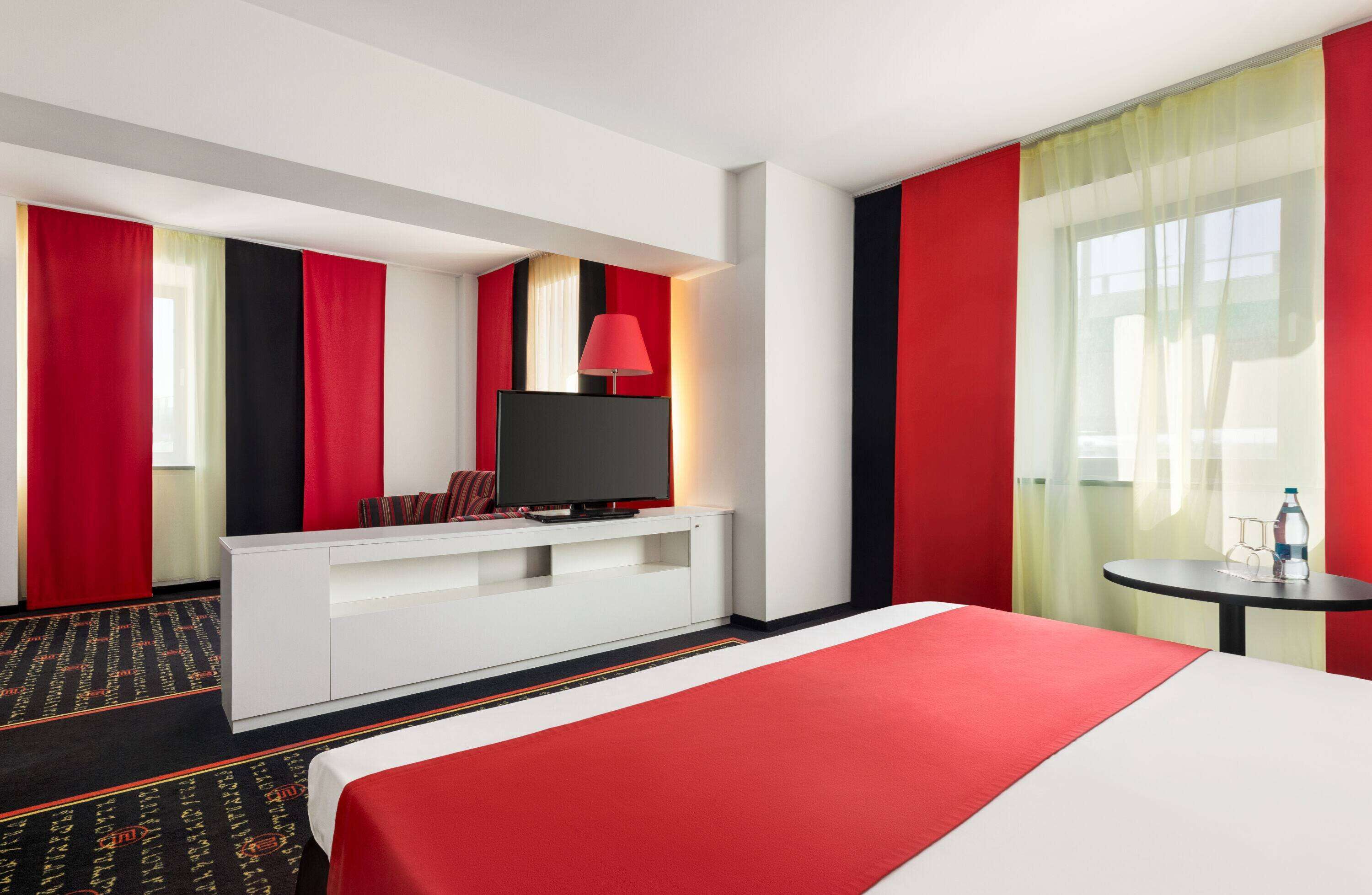 Ein Hotelzimmer mit einem Bett, einem Fernseher auf einer weißen Kommode, einem kleinen Tisch mit einer Flasche und roten Vorhängen.
