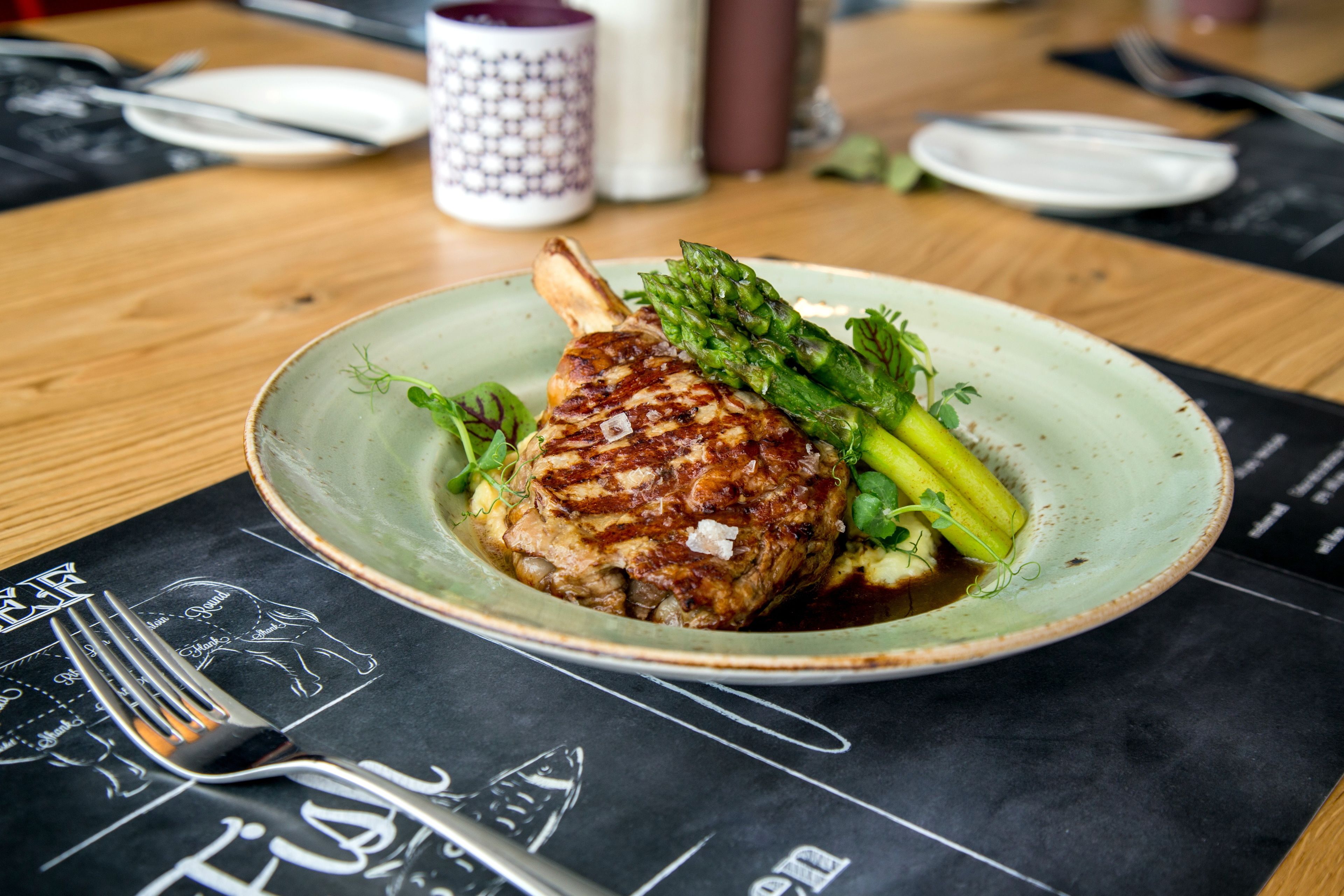 A plate with a grilled meat, asparagus, and a sauce on a table with a black placemat.