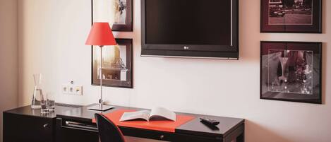 A hotel room with a desk, chair, TV, and framed pictures on the wall.