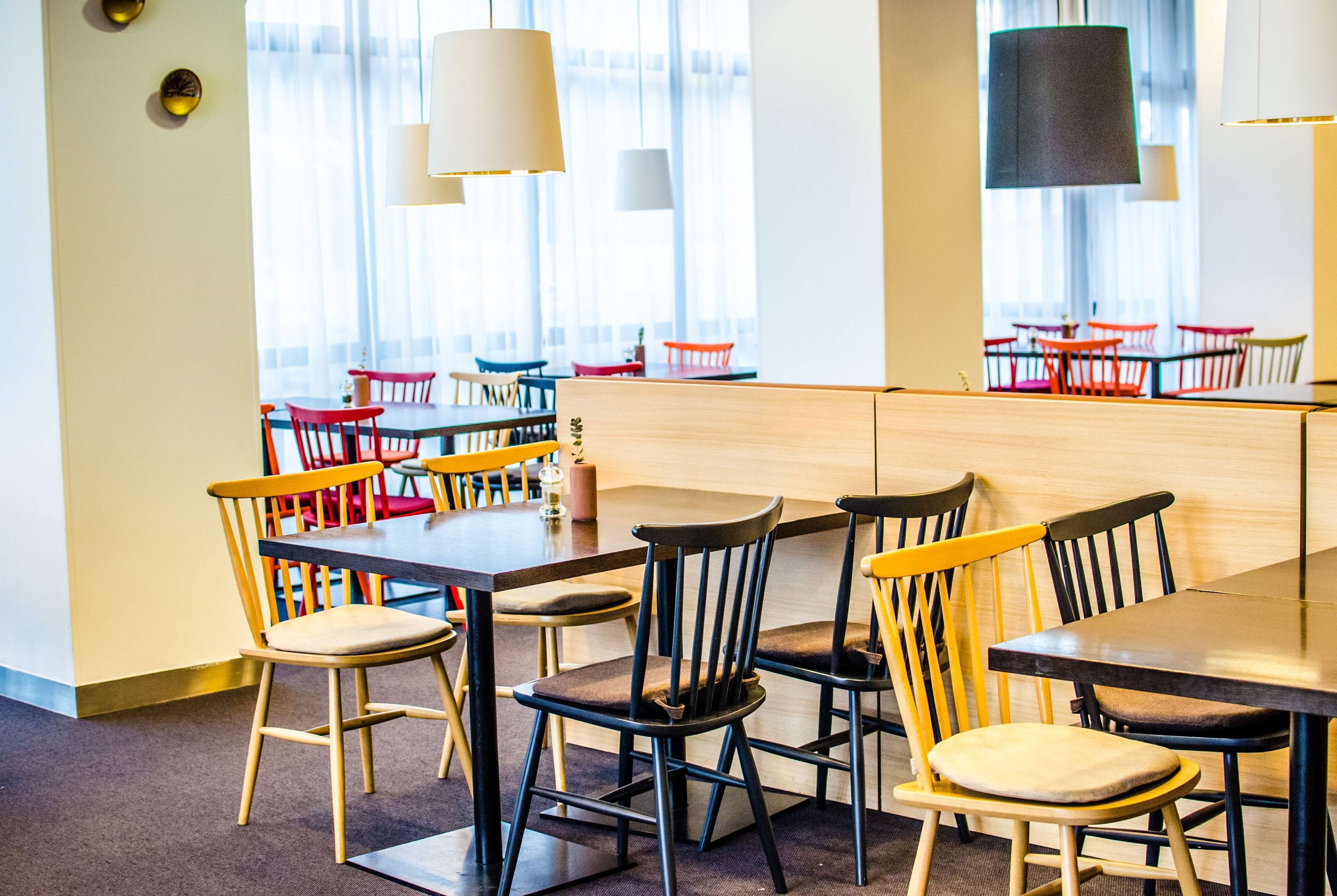 A dining area with multiple tables and chairs, large windows with curtains, and hanging pendant lights.