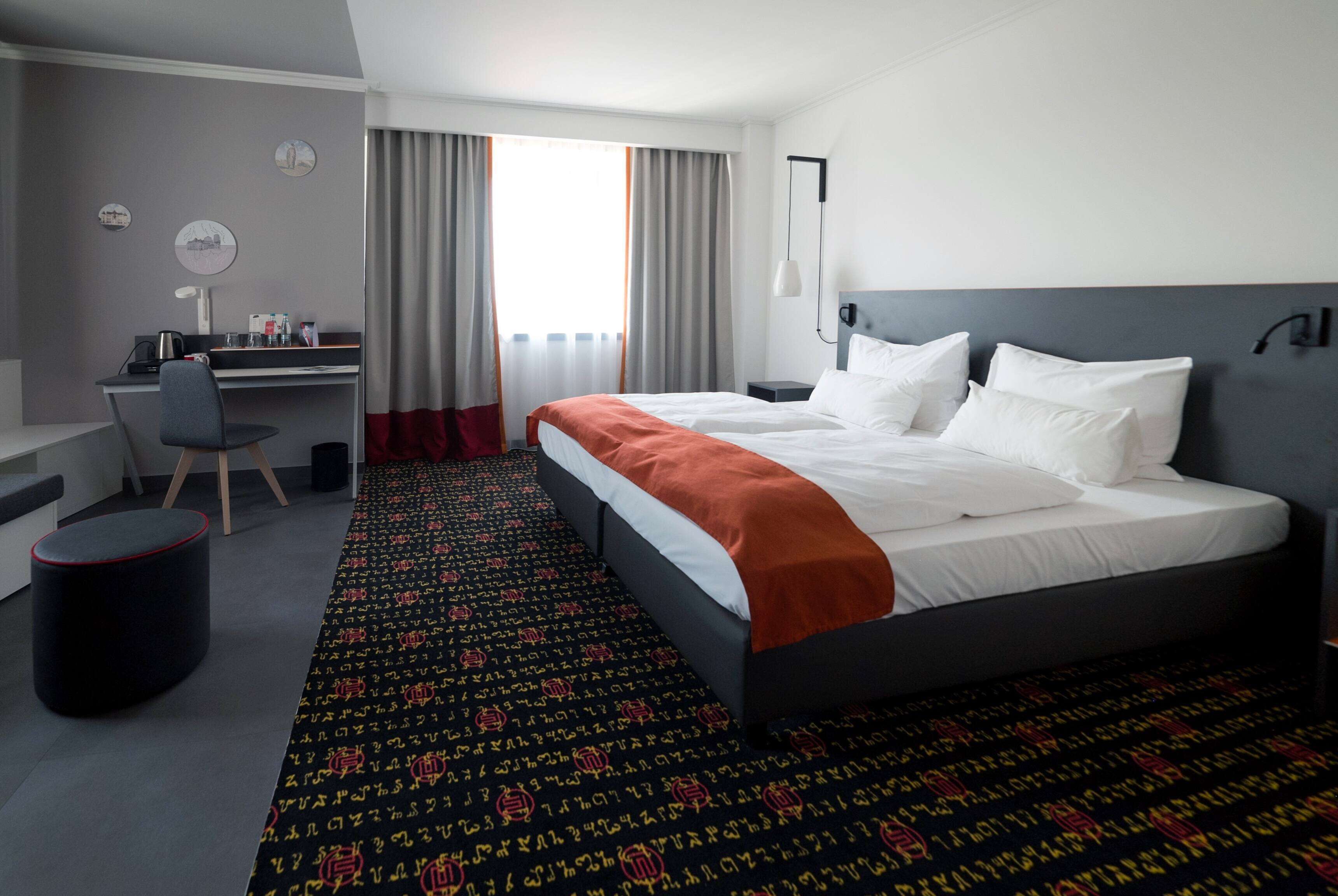 A hotel room with a bed, desk, chair, and a view of the city through the window.