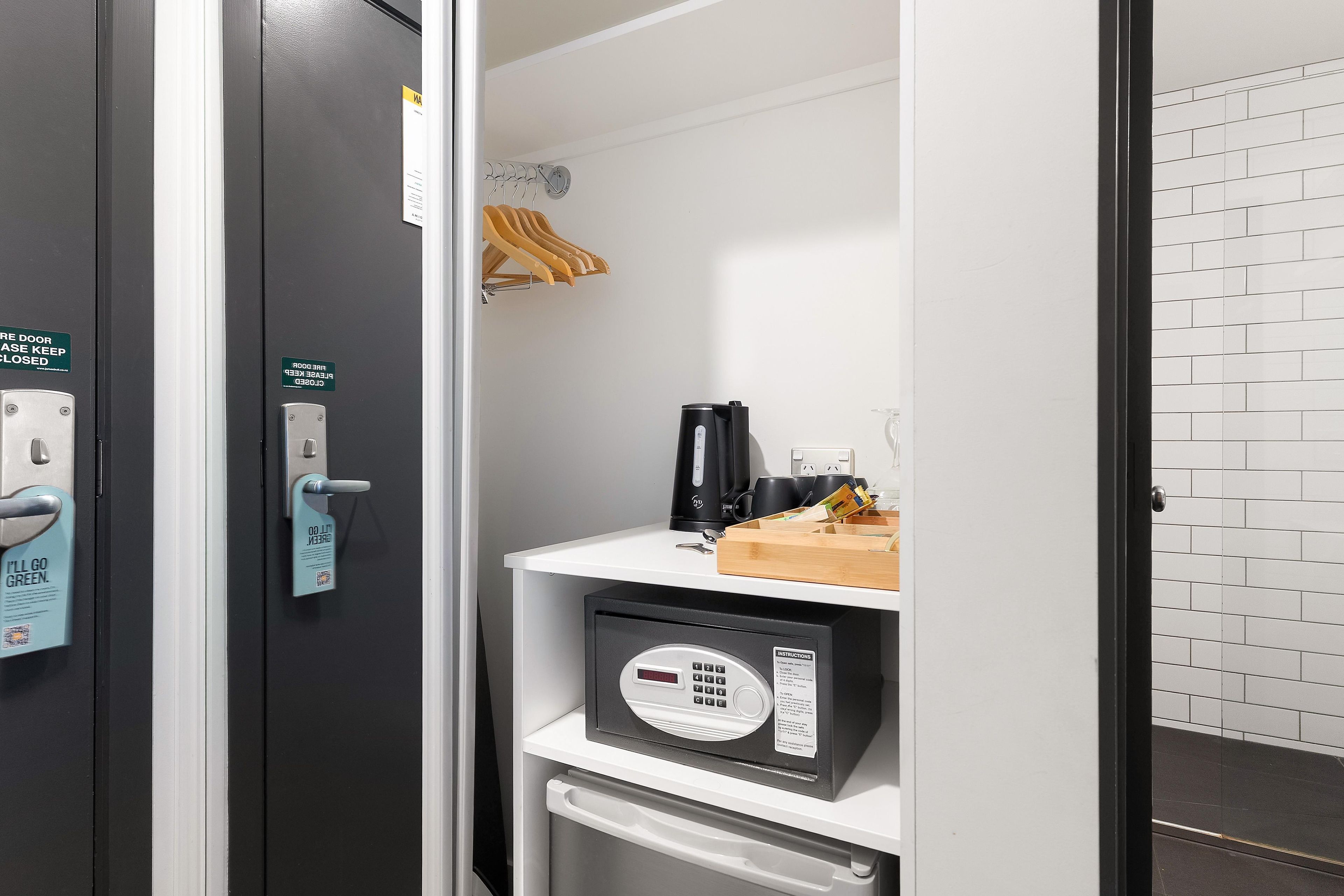 A hotel room with a door that has a green sign indicating it is closed, a keycard reader, a small refrigerator, a coffee machine, and a shelf with a wooden hanger.