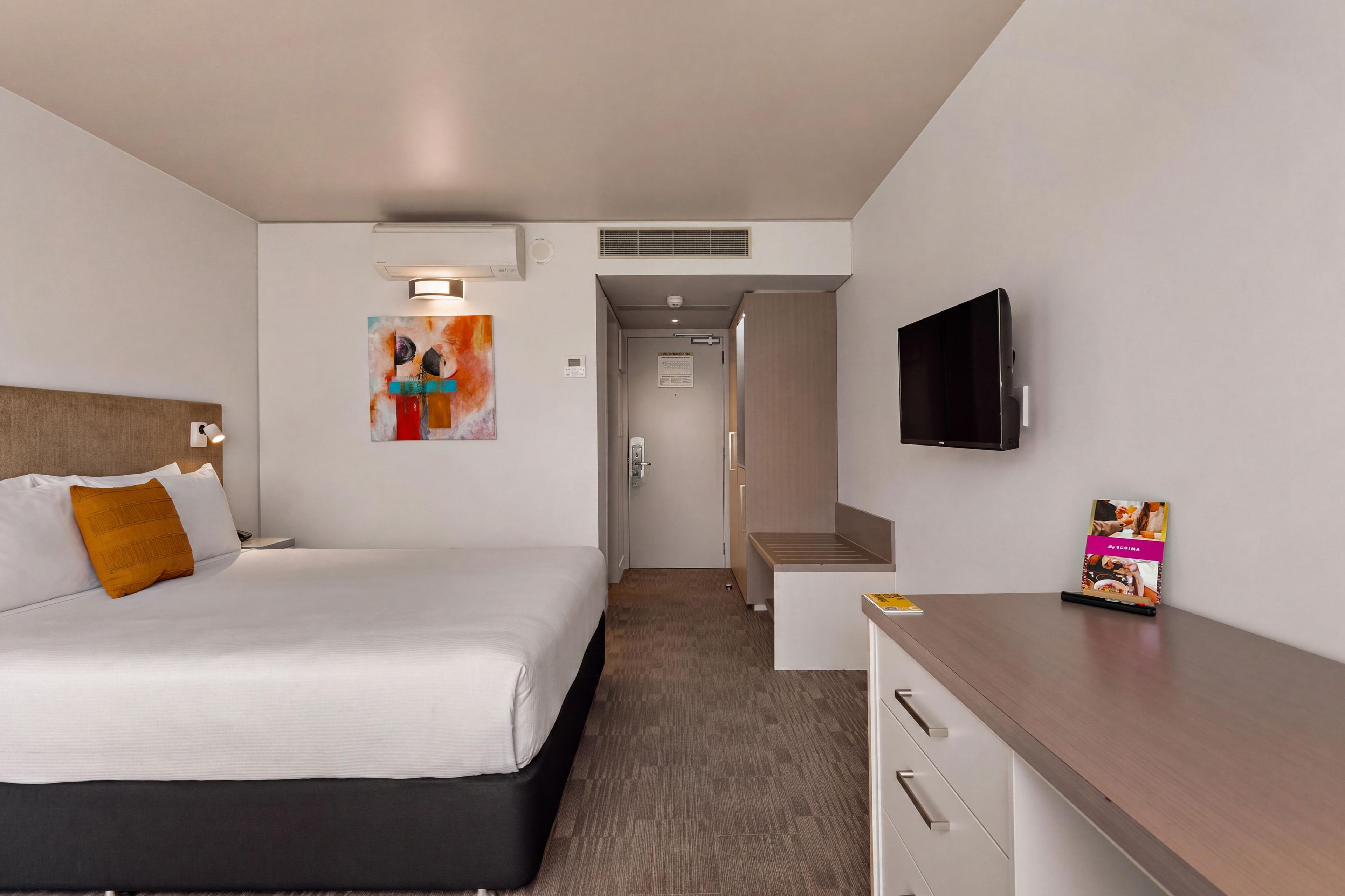A hotel room with a bed, a desk, a television, and a painting on the wall.