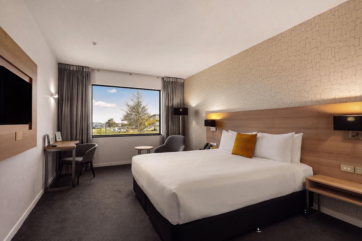 A hotel room with a large bed, a desk, a chair, a small table, a TV, and a window with a view of trees and buildings.