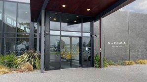 La entrada de Sudima Hotels con puertas de vidrio, un muro de concreto y un camión de reparto al fondo.