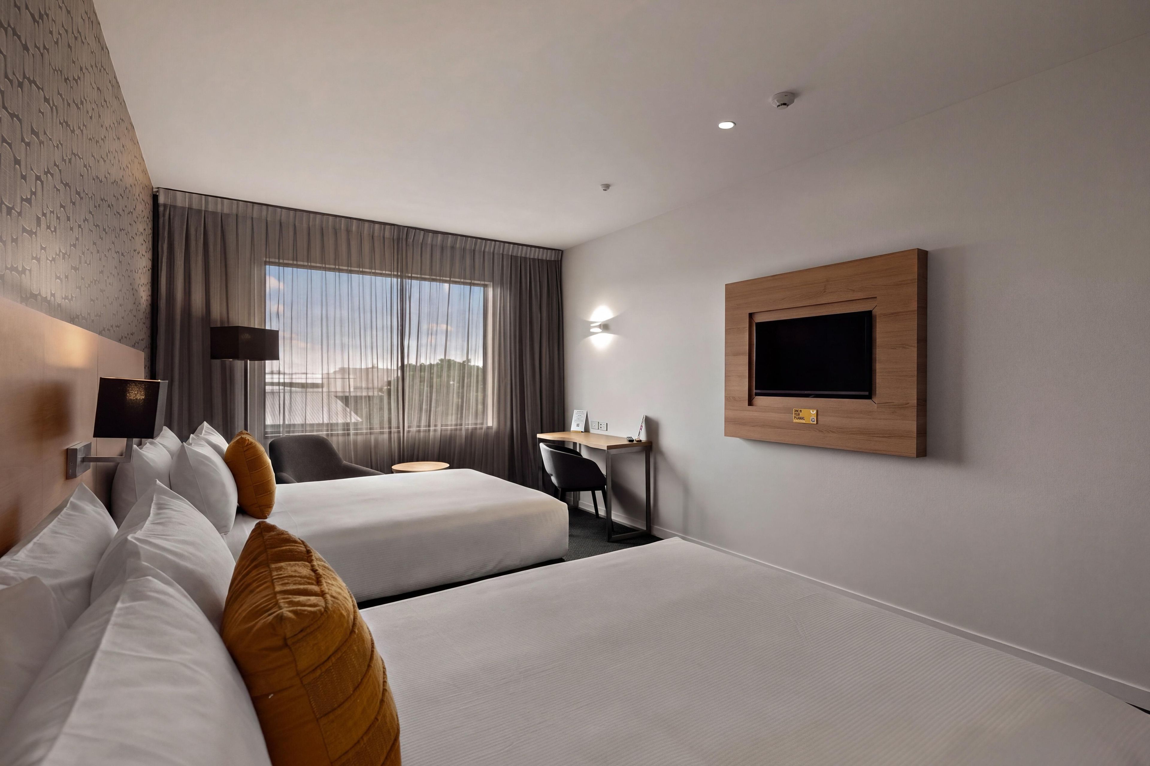 A hotel room with two beds, a desk, a chair, a TV mounted on the wall, and a window with curtains.