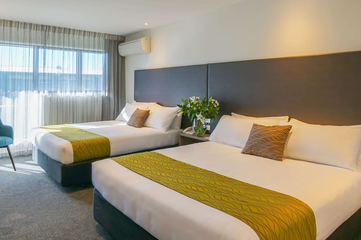A hotel room with two beds, a bedside table with a vase of flowers, and a view of the outdoors through large windows.