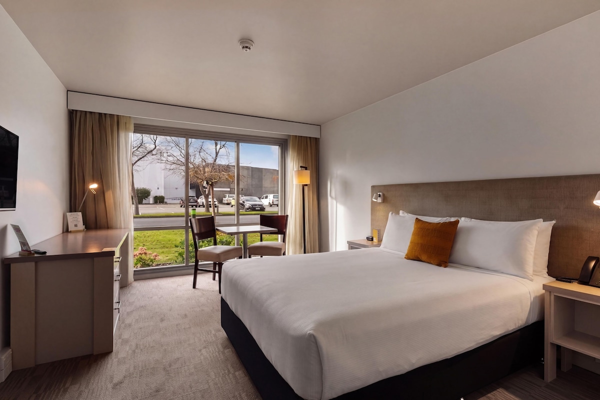 A hotel room with a large bed, a desk, and a chair. There is a window with a view of the street and cars.