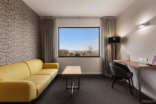 A hotel room with a yellow sofa, a small table, a desk with a chair, a large window with a view, and a wall with a leaf pattern.