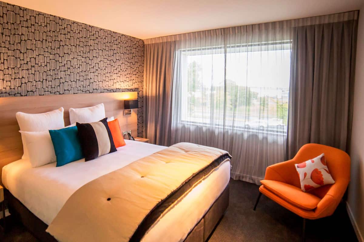 A hotel room with a large bed, a chair, a patterned wall, and a window with curtains.