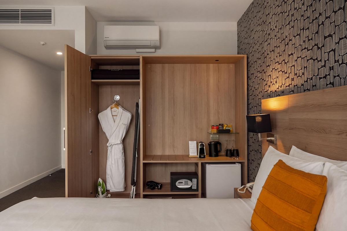 A hotel room with a bed, a wardrobe, a small kitchenette, and a wall-mounted TV.