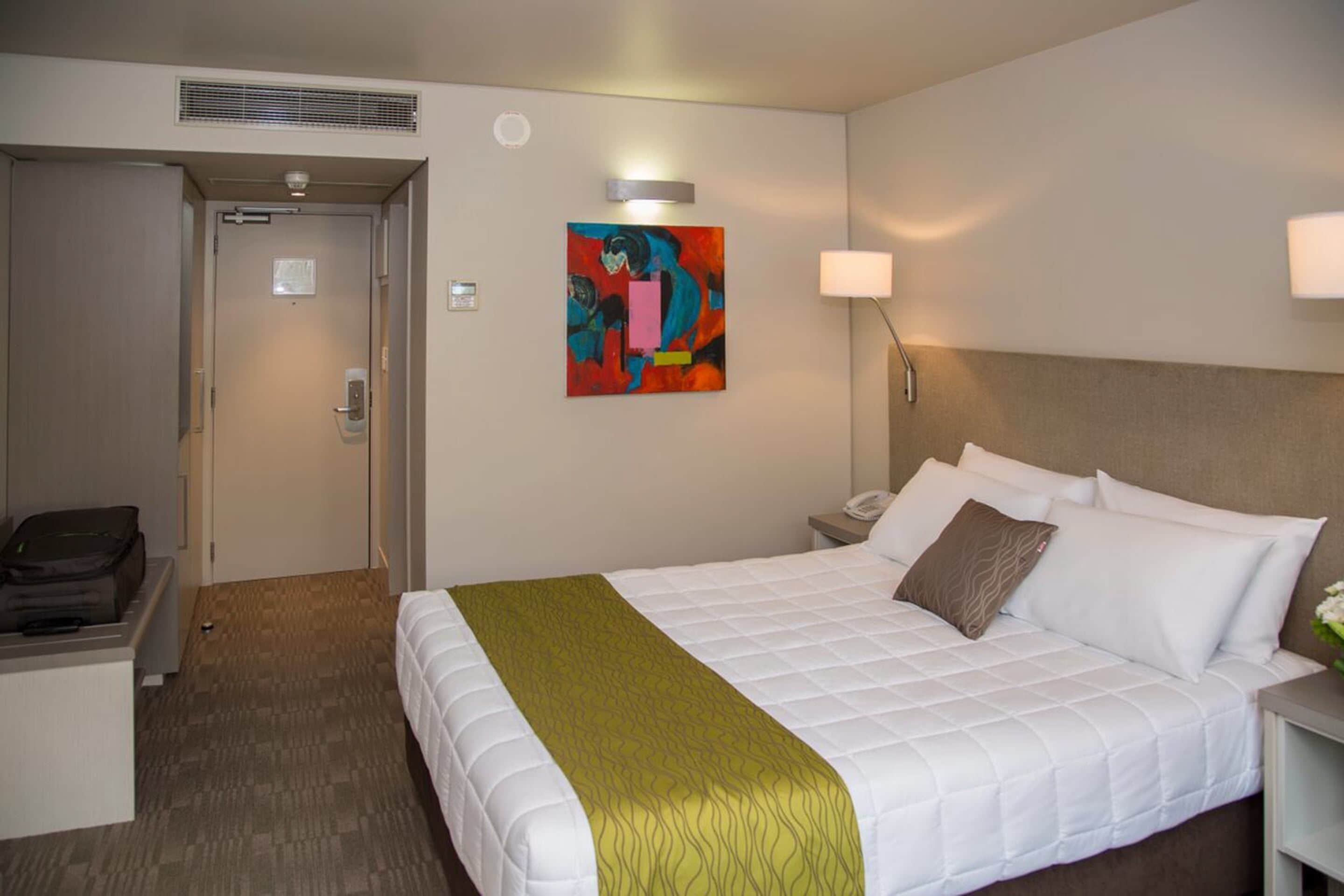 Una habitación de hotel con cama, mesita de noche, lámpara, una obra de arte en la pared y una puerta.