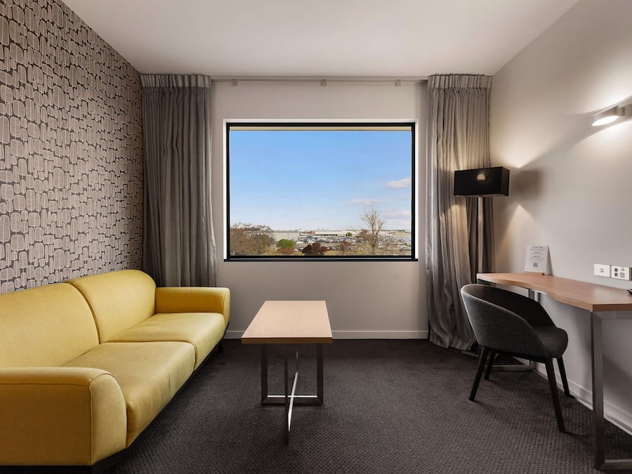 Habitación de hotel con sofá amarillo, una mesita, un escritorio con silla, un ventanal con vistas y una pared con diseño de hojas.
