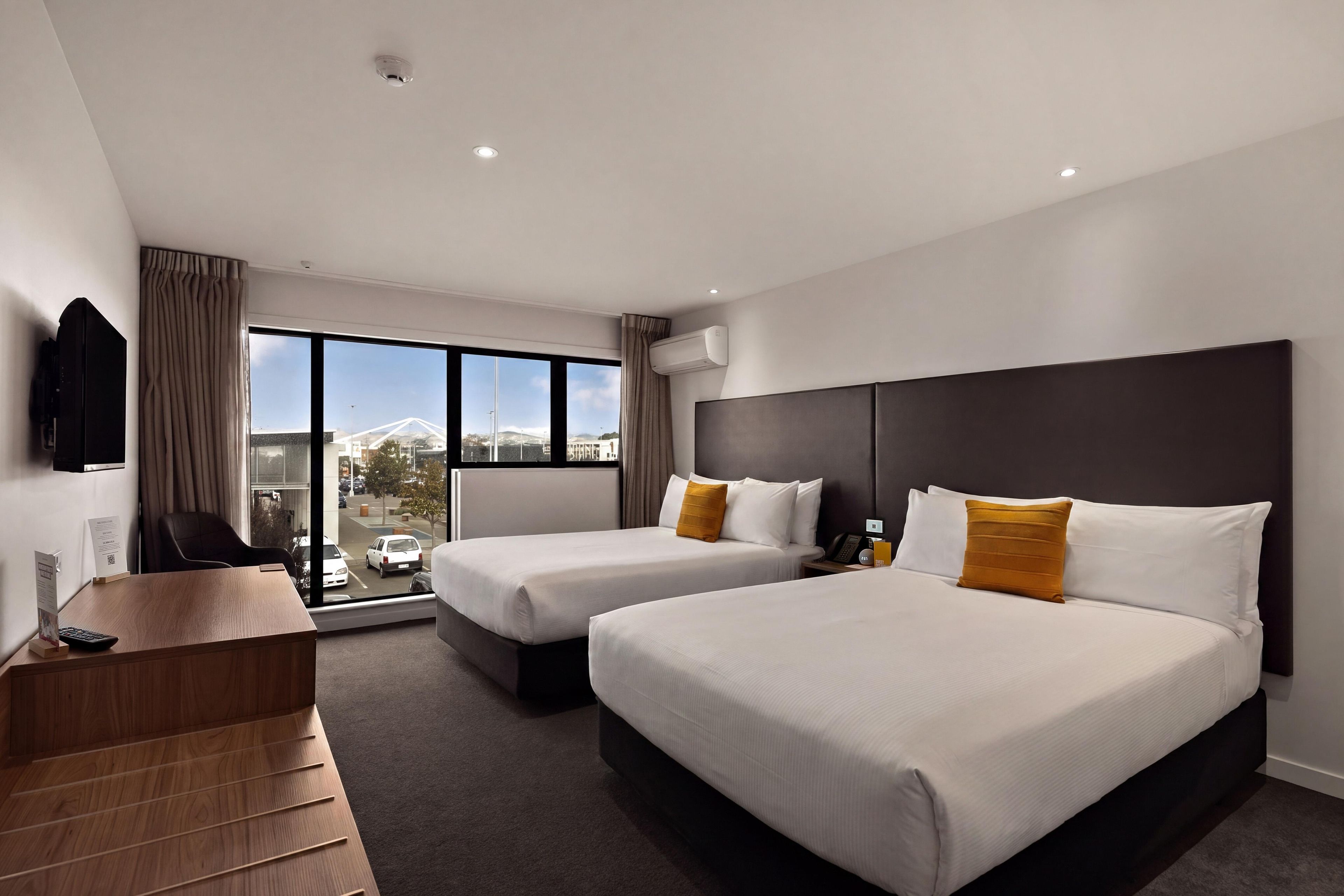 A hotel room with two beds, a desk, a chair, a TV, and a window with a view of the city.