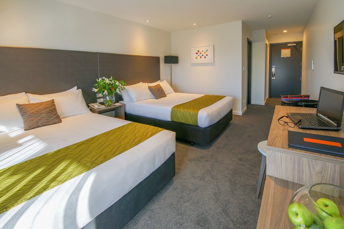 A hotel room with two beds, a desk with a laptop, and a vase with green apples.