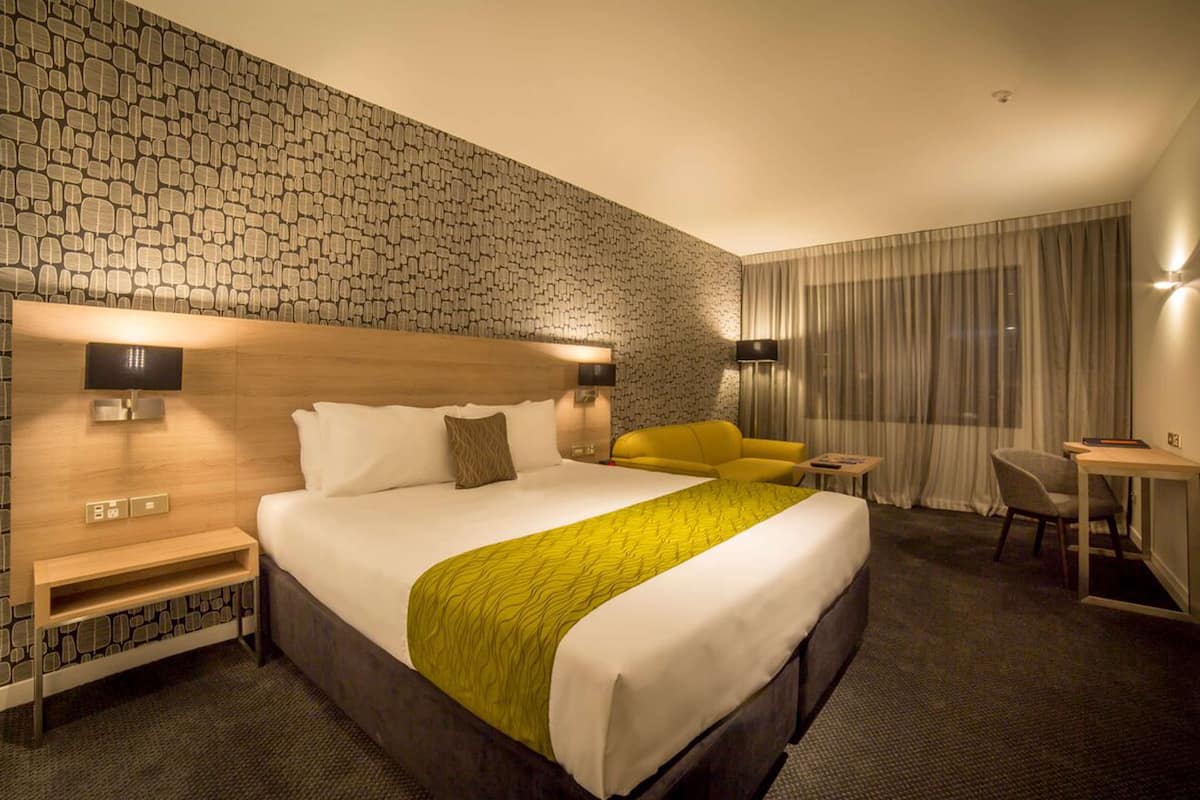 A hotel room with a large bed, a wooden headboard, a yellow armchair, a desk, and a small table.