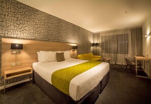 A hotel room with a large bed, a wooden headboard, a yellow armchair, a desk, and a small table.