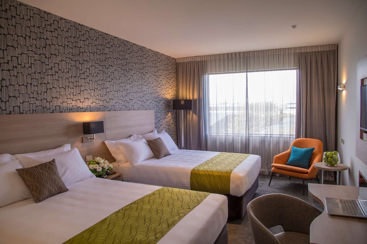 A hotel room with two beds, a small table with a laptop, a chair, and a large window with curtains.