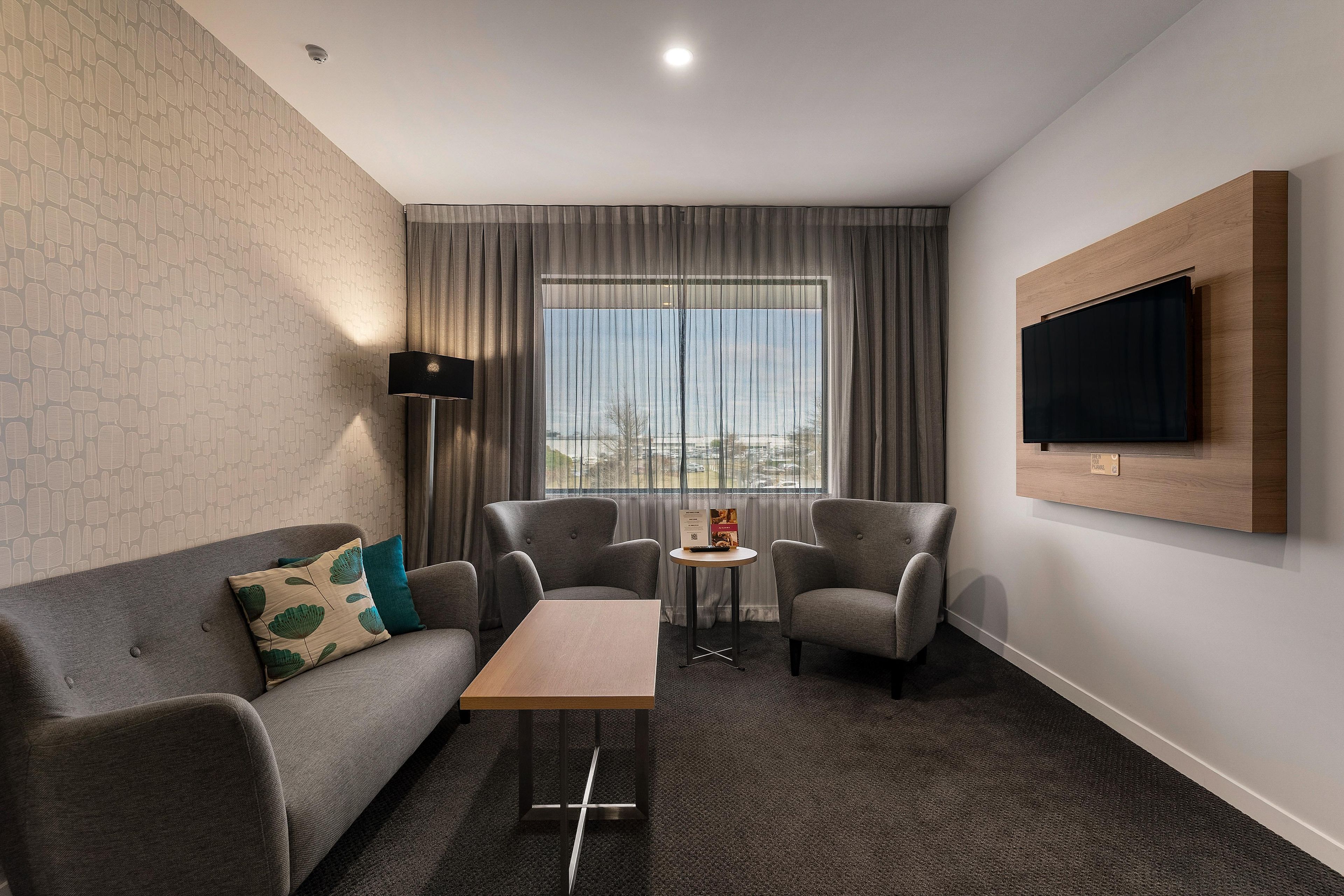 Una habitación de hotel moderna con un sofá gris, dos sillones, una pequeña mesa de centro de madera y un televisor de pantalla plana montado en la pared.