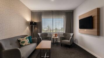 Una habitación de hotel moderna con un sofá gris, dos sillones, una pequeña mesa de centro de madera y un televisor de pantalla plana montado en la pared.