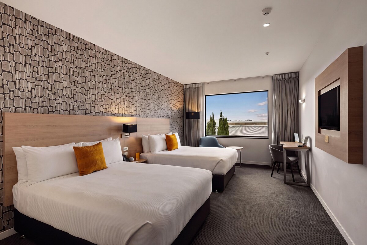 A hotel room with two beds, a desk, a chair, and a large window offering a view of the cityscape.