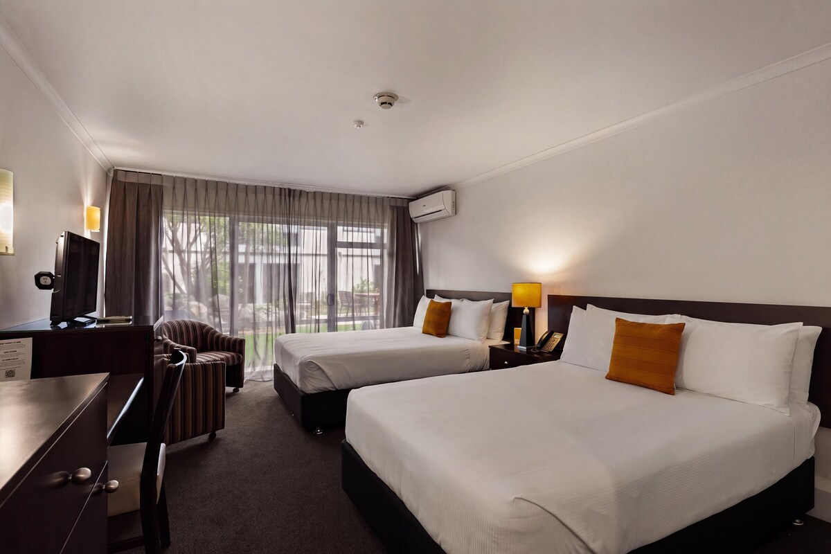 A hotel room with two beds, a desk, a chair, a TV, and an air conditioning unit.
