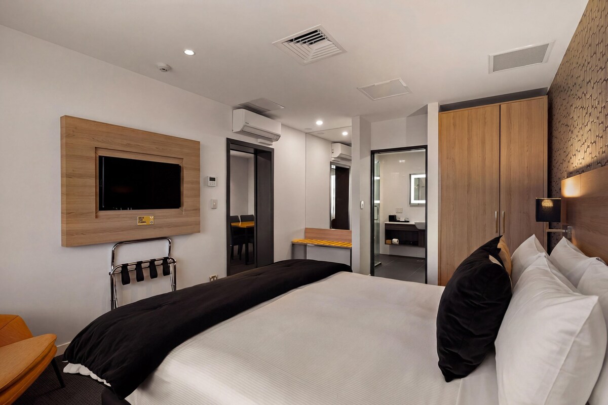 A modern hotel room with a large bed, a flat-screen TV mounted on the wall, a small table, and a wardrobe.