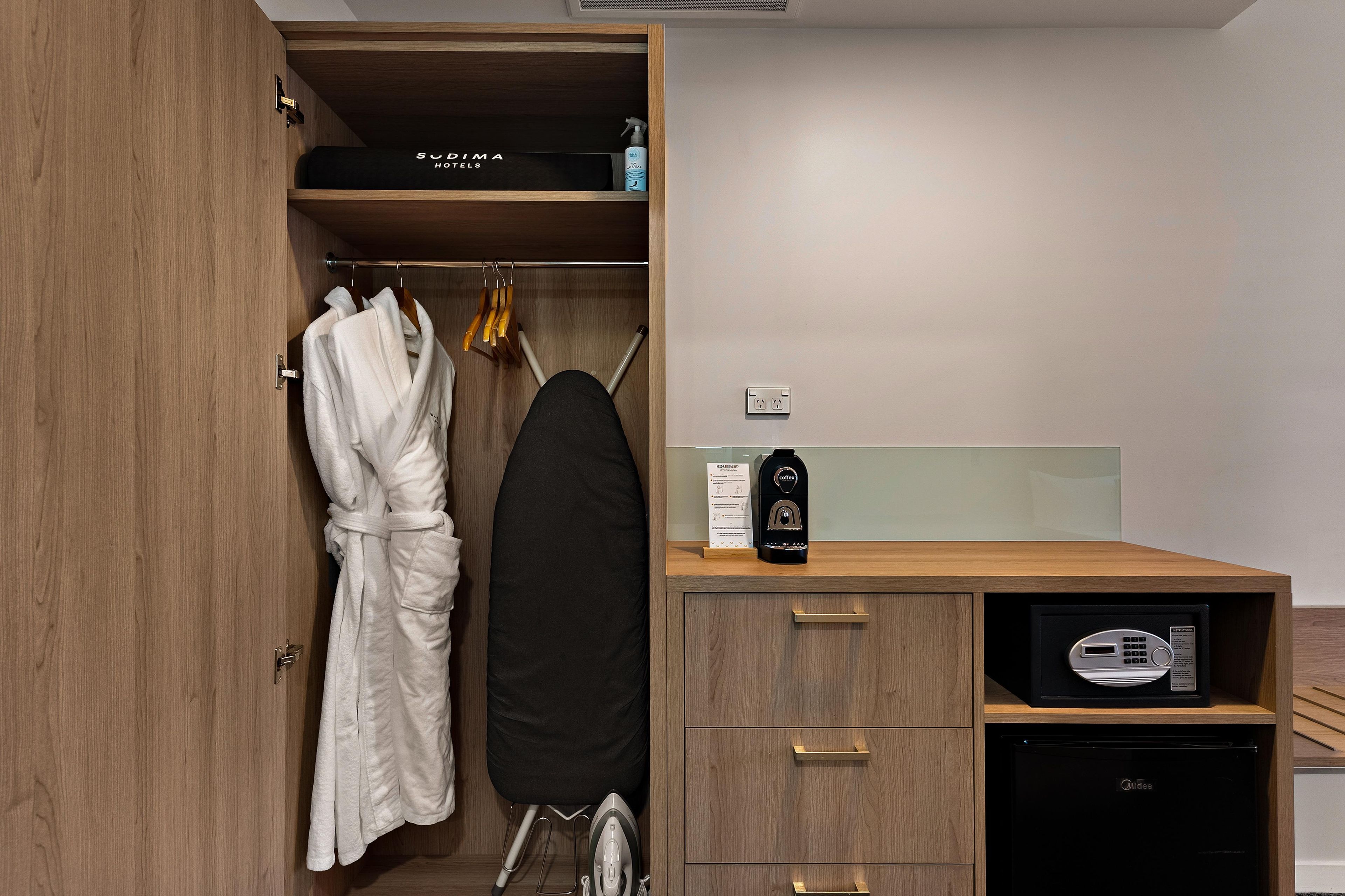 A hotel room with a wooden wardrobe, a robe hanging inside, a coffee machine, and a compact dishwasher.