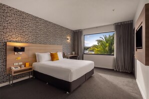 A hotel room with a large bed, a bedside table, a telephone, and a window with a view of palm trees. - Sudima Christchurch Airport (Christchurch)