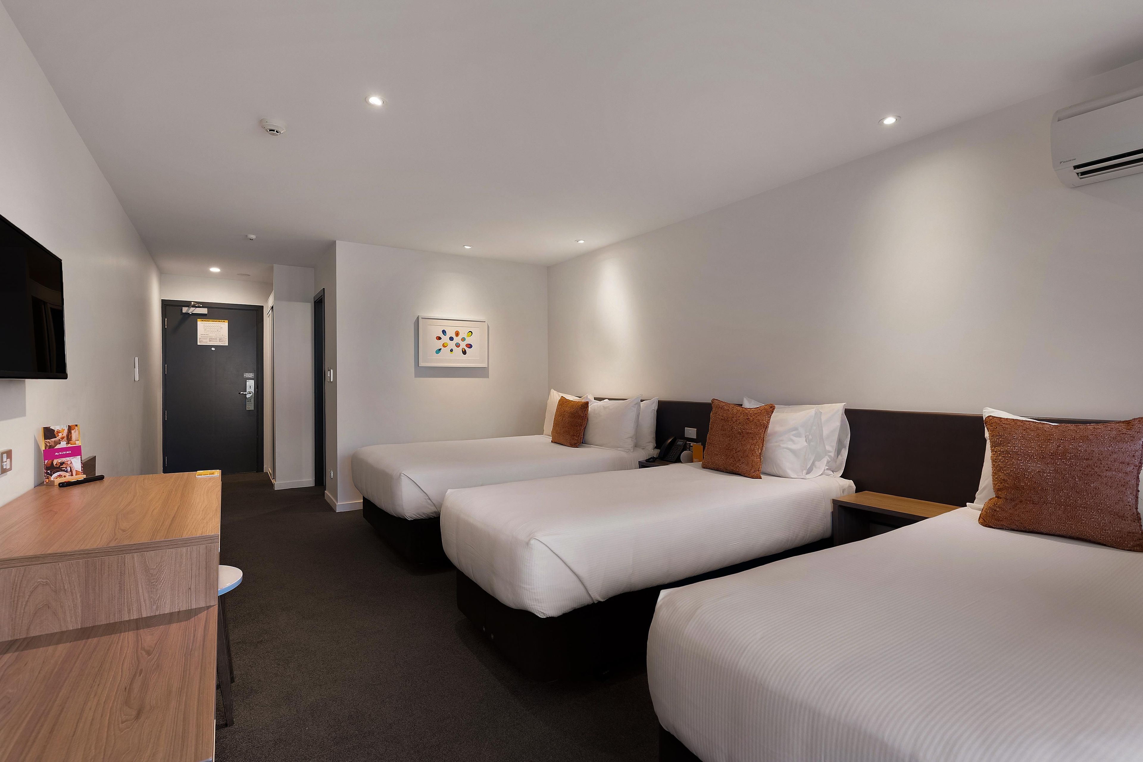 A hotel room with two beds, a desk, a flat-screen TV, and a wall-mounted air conditioner.