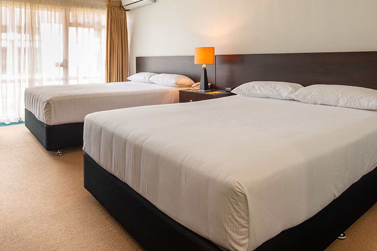 A hotel room with two beds, a nightstand with a lamp, and a window with curtains.